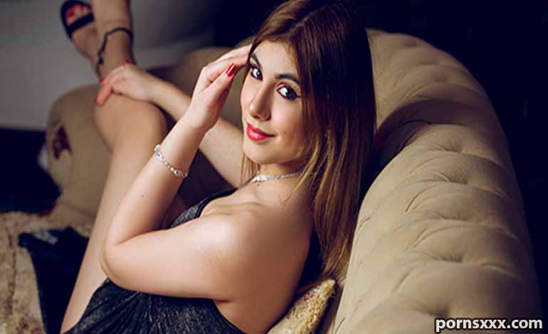 Gay Porn FM: Top Romantic Gifts to Surprise Your Partner Woman lounging on a beige couch, looking at the camera with a slight smile, wearing a dark dress and jewelry.