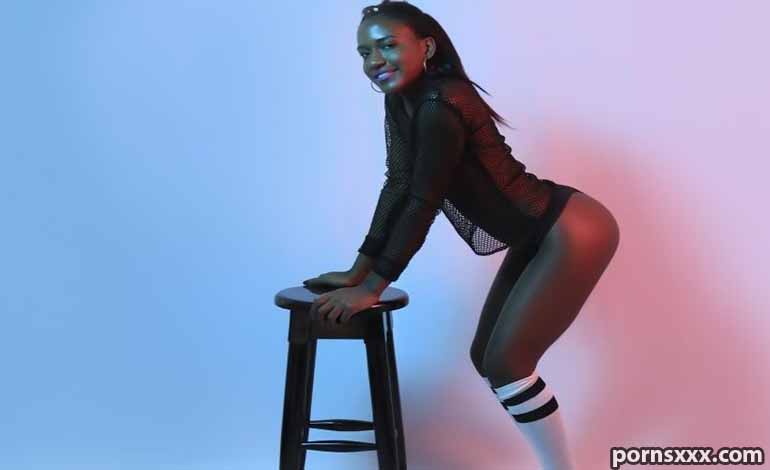 Royal Cams Sex: How Search Filters Help You Find The Best A woman in a black mesh bodysuit leans on a wooden stool in a studio with blue and pink gradient lighting, smiling at the camera.