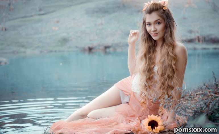 Shower Sex Toys Turn Your Daily Routine Into A Very Fun Escape Young woman with long wavy hair and a flower crown sits beside a lake in a peach dress, looking at the camera.